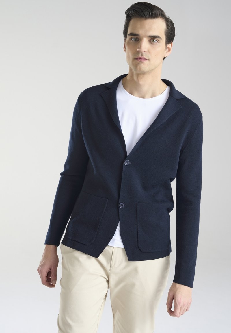 Granatowy kardigan z miękkiej dzianiny, z kołnierzem z wyłogami, dwiema przednimi kieszeniami i zapięciem na guziki, noszony na białej koszuli.