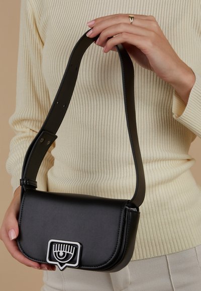 Sac bandoulière en cuir noir de forme incurvée, fermeture à boucle ornée d'un motif œil, et sangle plate noire. Surface texturée.