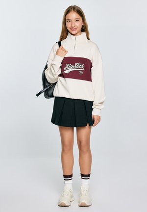 Sweatshirt zippé crème avec une rayure bordeaux portant le texte "Big Star 79", associé à une jupe plissée noire et des baskets beiges.