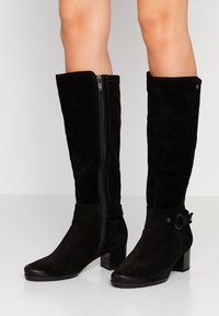 Botas de ante negras hasta la rodilla con cremallera lateral, una correa con un acento de anillo metálico y un tacón cuadrado para mayor estabilidad.