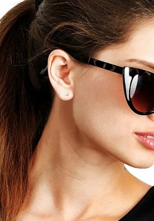 Lunettes de soleil noires et écaille de tortue avec des verres teintés foncés, portées avec une petite boucle d'oreille en perle, encadrées par des cheveux bruns raides en queue de cheval.