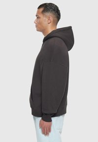 Dunkelgrauer Hoodie mit großer Kapuze, lockerem Schnitt, Fronttasche und gerippten Bündchen. Das Modell steht seitlich vor einem schlichten Hintergrund.
