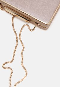 Käsilaukku, jossa on teksturoitu metallipinta ja kultainen metallikehys, kiinnitetty kultaketjuolkaimeen, valkoista taustaa vasten.