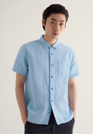 Giovane uomo con capelli scuri che indossa una camicia azzurra a maniche corte con taschino sul petto e pantaloni neri, su sfondo semplice.