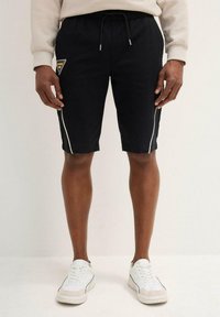 Pantaloni corti neri con strisce bianche ai lati, cintura elastica con coulisse, caratterizzati da una patch con logo a forma di triangolo sulla coscia sinistra.
