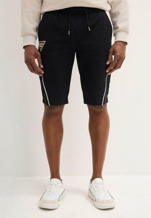 Shorts noirs avec des rayures latérales blanches, taille élastique avec cordon de serrage, présentant un patch logo en forme de triangle sur la cuisse gauche.
