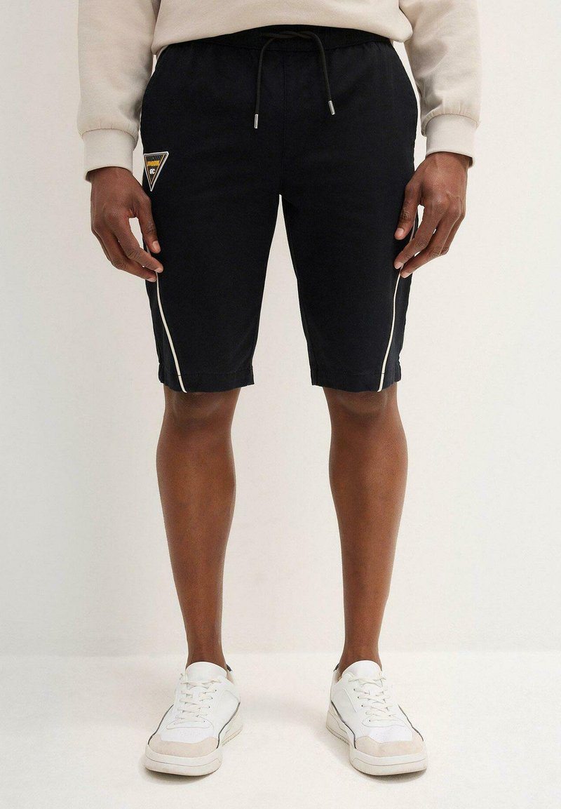 Pantaloni corti neri con strisce bianche ai lati, cintura elastica con coulisse, caratterizzati da una patch con logo a forma di triangolo sulla coscia sinistra.