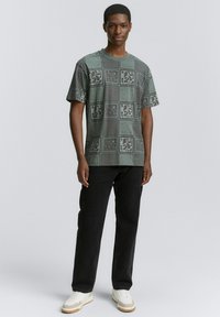 Grøn t-shirt med mønster, der har forskellige designs, kortærmet og rund halsudskæring, parret med sorte jeans og hvide sneakers.