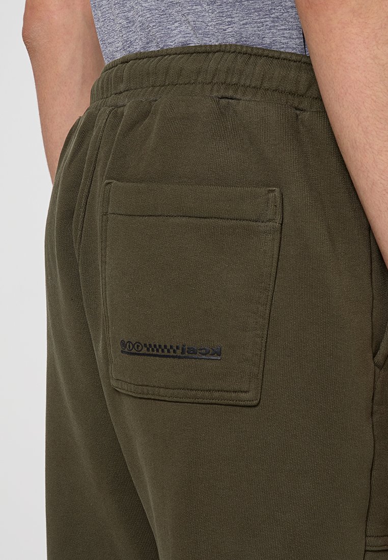 Joggers de color verde oliva, confeccionados con una tela suave, presentan un bolsillo trasero con costuras negras y un logo. La cinturilla es elástica para mayor comodidad.