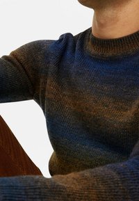 Homme portant un pull en tricot dégradé bleu et marron avec encolure et manches côtelées, assis avec un bras plié visible.