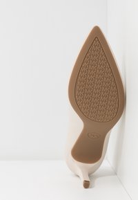 Chaussure à talon haut beige avec bout pointu, affichant une semelle embossée du logo MK répétitif et un petit talon.