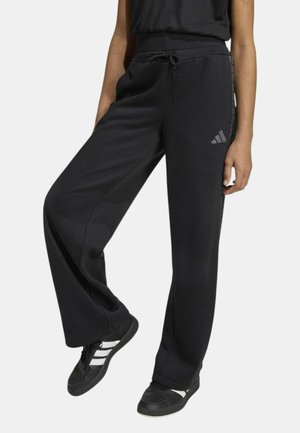 Persona che indossa pantaloni sportivi neri a gamba larga con vita a coulisse e scarpe da ginnastica nere con strisce bianche, in piedi contro uno sfondo semplice.