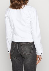 Mujer con una chaqueta de mezclilla blanca y jeans negros de tiro alto, vista desde atrás contra un fondo plano.