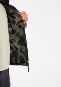Olijfgroene jacket met een zwarte voering die een abstract patroon heeft. Voorzien van een ritssluiting en elastische manchetten. Lichtgewicht materiaal.