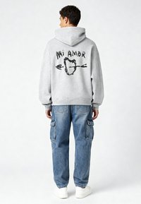 Pilkas džemperis su suvarstomu gobtuvu, turintis juodą grafišką dizainą ant nugaros. Suderintas su šviesiai mėlynomis cargo kelnėmis ir baltomis sportinėmis batu.