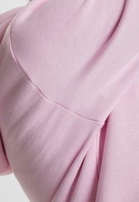 Tela de sudadera color rosa claro con una textura suave. Presenta una costura curva en el hombro y pliegues en el lateral para un detalle adicional.