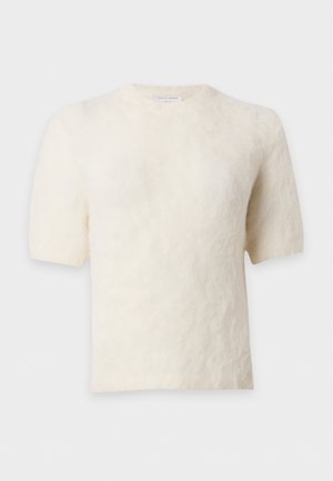 Un mannequin se tient devant un fond de studio blanc, vêtu d’un pull crème à manches courtes et d’un pantalon large bleu marine, avec un sac en cuir noir à la main.