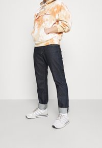 Sweatshirt i tie-dye orange och krämfärgad, stor framficka; mörk denimjeans med upprullade fållar; vita sneakers med grå detaljer.