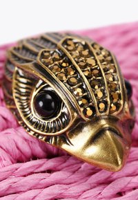 Bronzowy brosz w kształcie żaby z czarnymi kryształowymi oczami i brązowymi cyrkoniami, spoczywający na teksturowanym, różowym plecionym materiale.