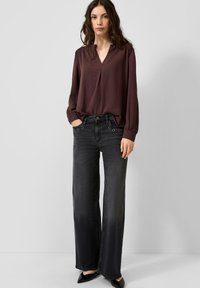 Bordeauxrode blouse met lange mouwen, een V-hals en geplooide schouders, gecombineerd met zwart denim jeans met wijde pijpen en cirkelvormige metalen details.