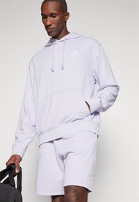 Felpa viola chiaro con cappuccio, con lacci e tasca a marsupio, abbinata a pantaloni corti coordinati. Il materiale appare morbido e informale.
