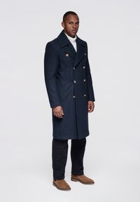 Marineblå dobbeltradet uldfrakke med guldknapper, hakket krave og sidelommer, kombineret med sorte bukser og brune ankelstøvler.