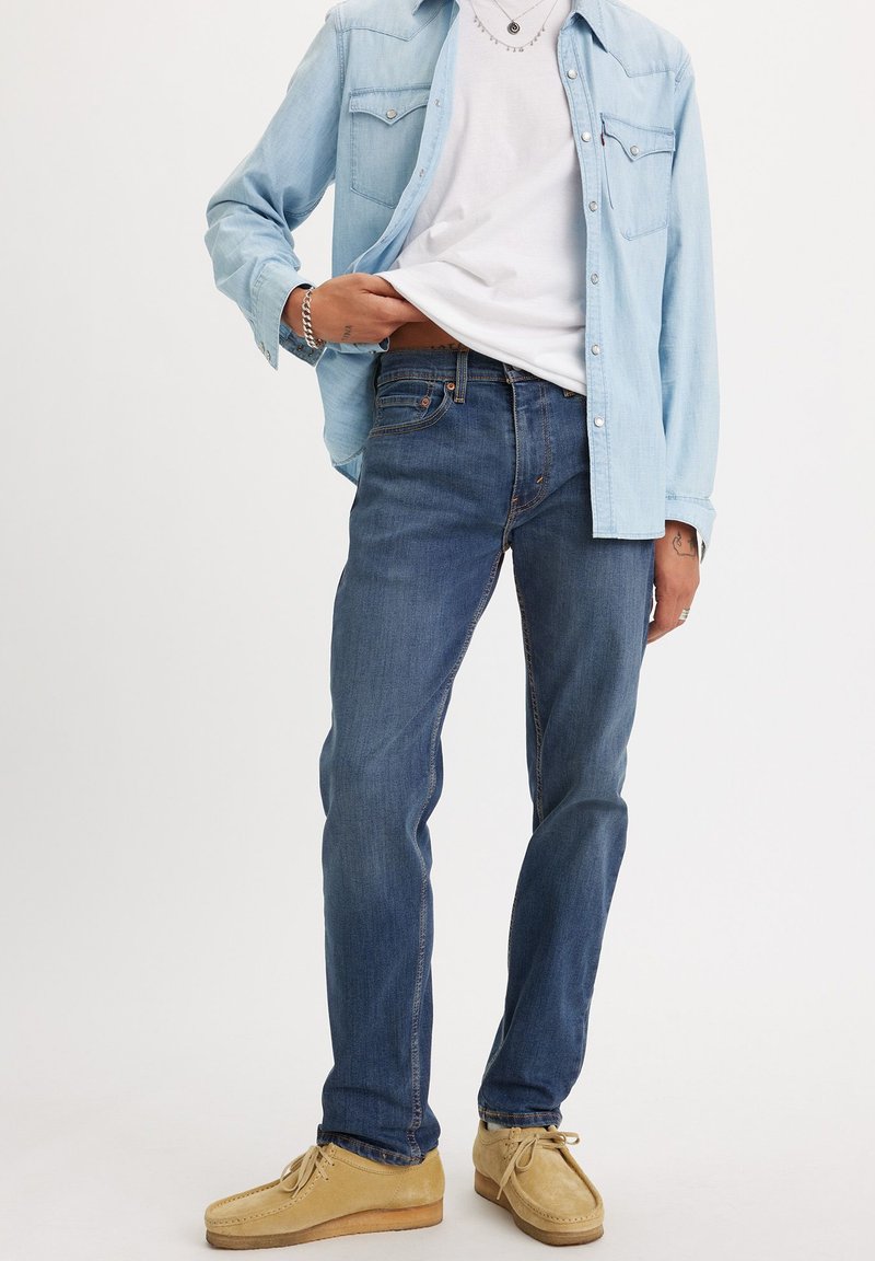 Hellblaues Jeanshemd, das über einem weißen T-Shirt getragen wird, kombiniert mit dunkelblauen Jeans und beigen Schnürschuhen. Gelegentliche Nähedetails sichtbar.