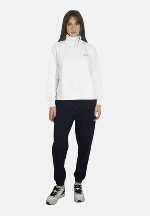 Felpa bianca a collo alto con logo ricamato, abbinata a pantaloni jogger blu scuro. Il modello indossa sneakers bianche con accenti neri e verdi.