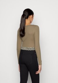 Kvinna med lång mörk hästsvans, klädd i en olivgrön långärmad topp med spetsdetaljer och högmidjade svarta jeans, stående med ryggen mot kameran.