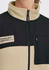 Fleece jack met een zwarte bovenkant en beige onderkant, met een hoge kraag, voorde rits en een borstzak met ritsdetail. Etiket inbegrepen.