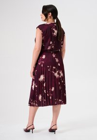 Pflaumenblumenkleid mit kurzen Ärmeln, gefaltetem Rock und einem Taillengürtel. Glatte, glänzende Stoffqualität, kombiniert mit offenen High Heels.