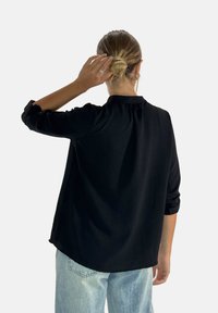 Maglietta nera a maniche lunghe con vestibilità rilassata, caratterizzata da un dettaglio arricciato sul retro e polsini arrotolati, abbinata a jeans di denim azzurro chiaro.