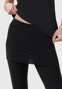 Zwart nauwsluitend topje over een zwarte rok gedragen, waarbij een deel van de tailleband zichtbaar is. De stof is glad en rekbaar.