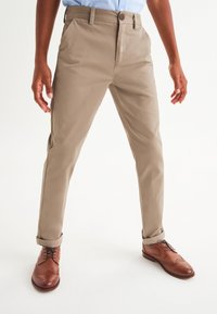 Beige Baumwollhosen mit geradem Schnitt, ausgestattet mit einem Knopfverschluss, Seitentaschen und aufgerollten Bündchen. Braune Lederschuhe mit Brogue-Details.