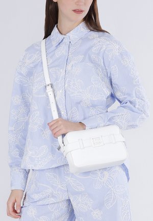 Vrouw draagt lichtblauw shirt en broek met bloemmotief, houdt een witte rechthoekige crossbodytas met verstelbare band vast.