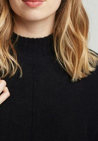 Pull en tricot noir avec un col côtelé. Texture douce, couture minimale visible, associé à des cheveux légers et ondulés au premier plan.