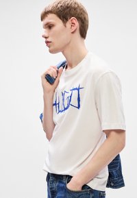 Tricou alb din bumbac cu un logo grafic albastru "HUGO." Guler rotund, mâneci scurte. Modelul ține o geacă din denim peste umărul unuia.