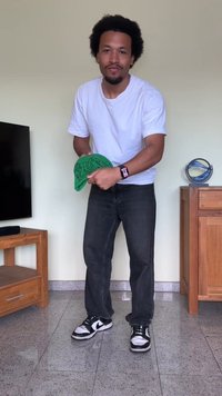 Camiseta branca, jeans escuros e ténis pretos com detalhes brancos. Boné verde com um design gráfico segurado nas mãos. Ambiente interior simples.