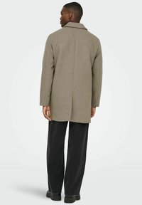 Olive roheline villasegu mantel, millel on sooniline krae, sirge lõige ja nähtavad õmblused, kombineeritud mustade laia säärega pükstega.