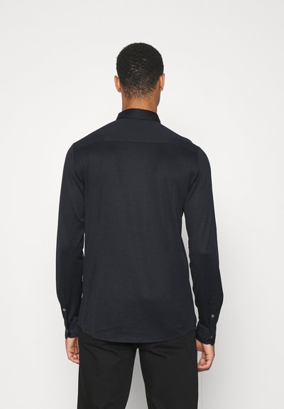 Maglietta a maniche lunghe blu navy scuro con colletto, texture liscia, polsini con bottoni e orlo curvo, vista dal retro. Design semplice e minimalista.