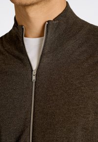 Maglione zip del colore marrone scuro con colletto alto e texture a coste. Presenta una cerniera argento sul davanti e una camicia bianca visibile sotto.