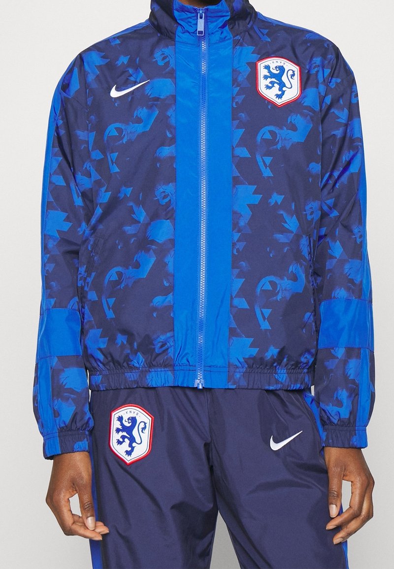 Blå och marinblå mönstrad jacka med dragkedja, axelpatchar och ett lejonemblem. Tillverkad av lätt material och med elastiska muddar.