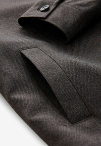 Brown textured fabric with a subtle striped pattern, featuring a buttoned cuff and a pocket detail. Focus on tailored fit and material quality.