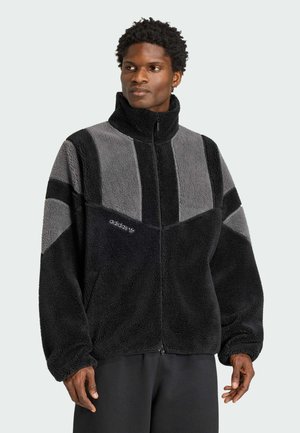 Man draagt een zwart-grijze fleece jas met rits en hoge kraag met het adidas-logo op de borst, staand tegen een effen achtergrond.