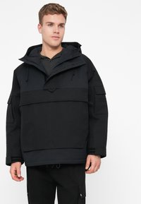 Casaco isolado preto com capuz, apresentando um grande bolso frontal, cordões ajustáveis e vários bolsos de utilidade nas mangas.