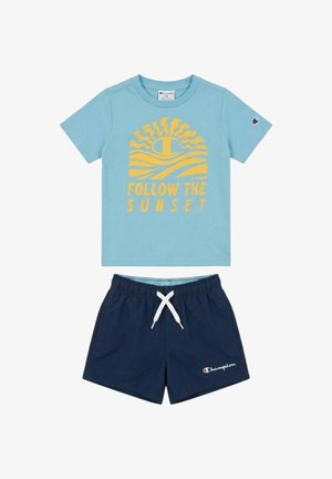 Camicia azzurro chiaro a maniche corte con grafica di sole giallo e testo "Follow the Sunset", abbinata a pantaloncini blu navy con coulisse bianca.