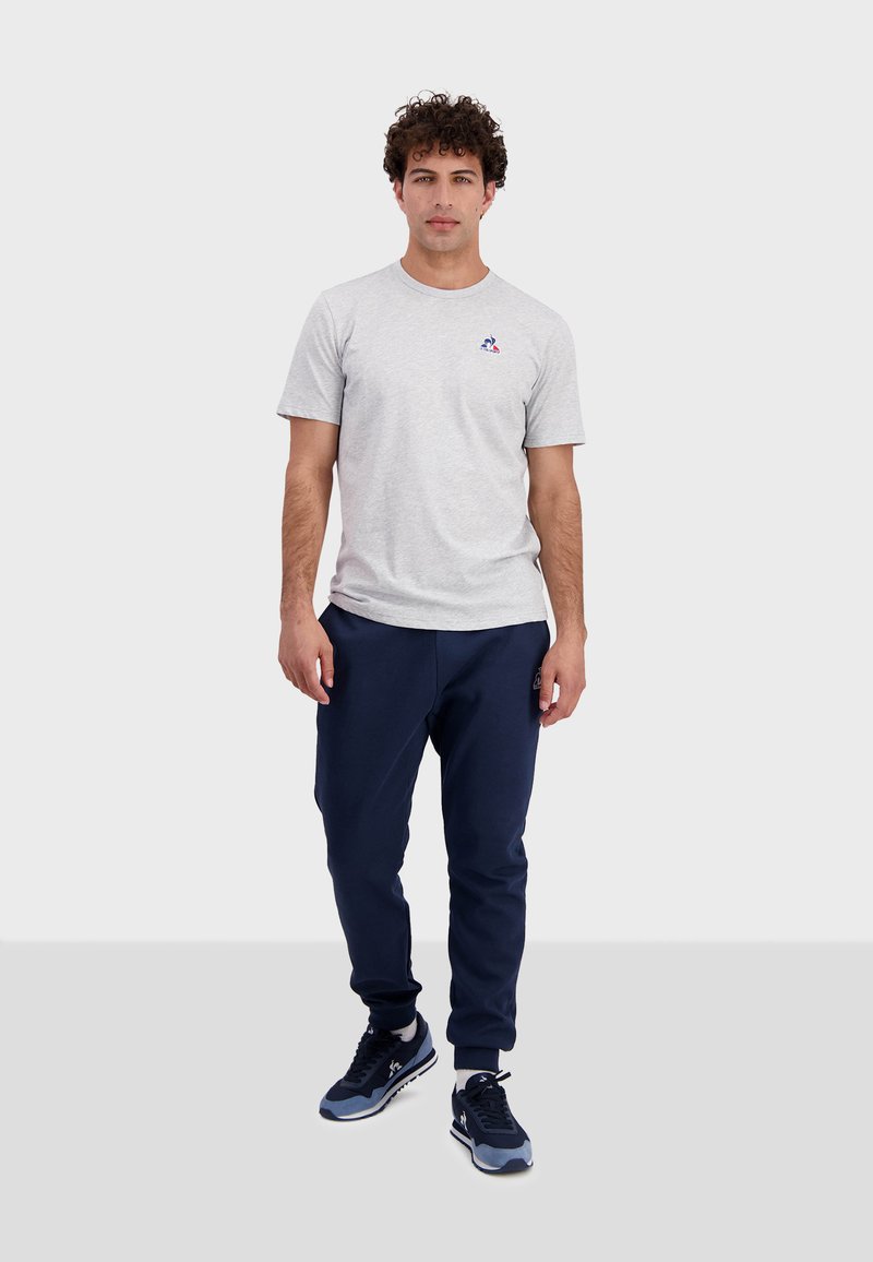 Grijze t-shirt met korte mouwen en een klein logo op de borst, gecombineerd met marineblauwe sweatpants en donkere sneakers, staand op een effen achtergrond.