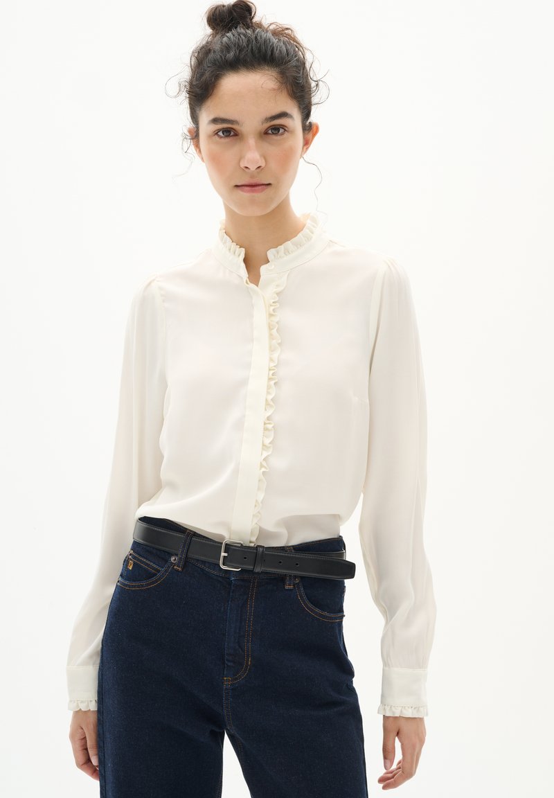 Blusa blanca con cuello de volantes y botones al frente, con mangas largas y una textura suave, combinada con jeans azul oscuro y un cinturón negro.