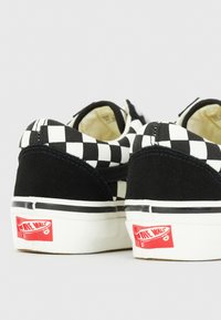 Zapatillas de lona en blanco y negro con diseño de tablero de ajedrez, detalles en ante negro, cuello acolchado, suela de goma y logo rojo "Off the Wall" en el talón.
