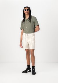 Olivengrøn kortærmet skjorte med knapper, beige stribede shorts med snoretræk, sorte sneakers, runde solbriller, glat tekstur.